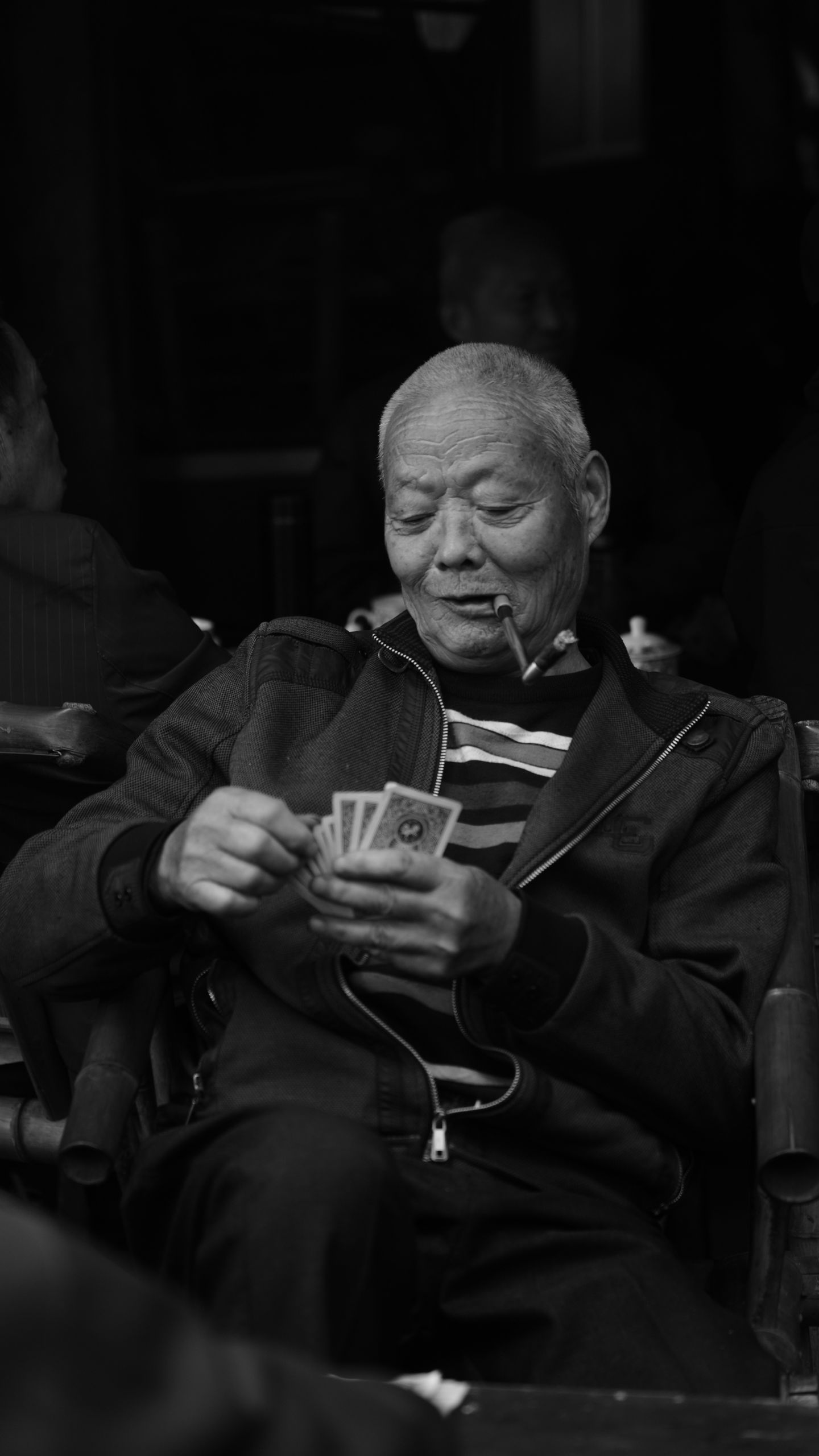 Black and white photo of a senior man enjoying a card game while smoking, capturing candid leisure moments.